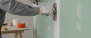 Plasterer smoothing drywall tape for plasterboard joints onto a pale green wall using a metal trowel and white gloves